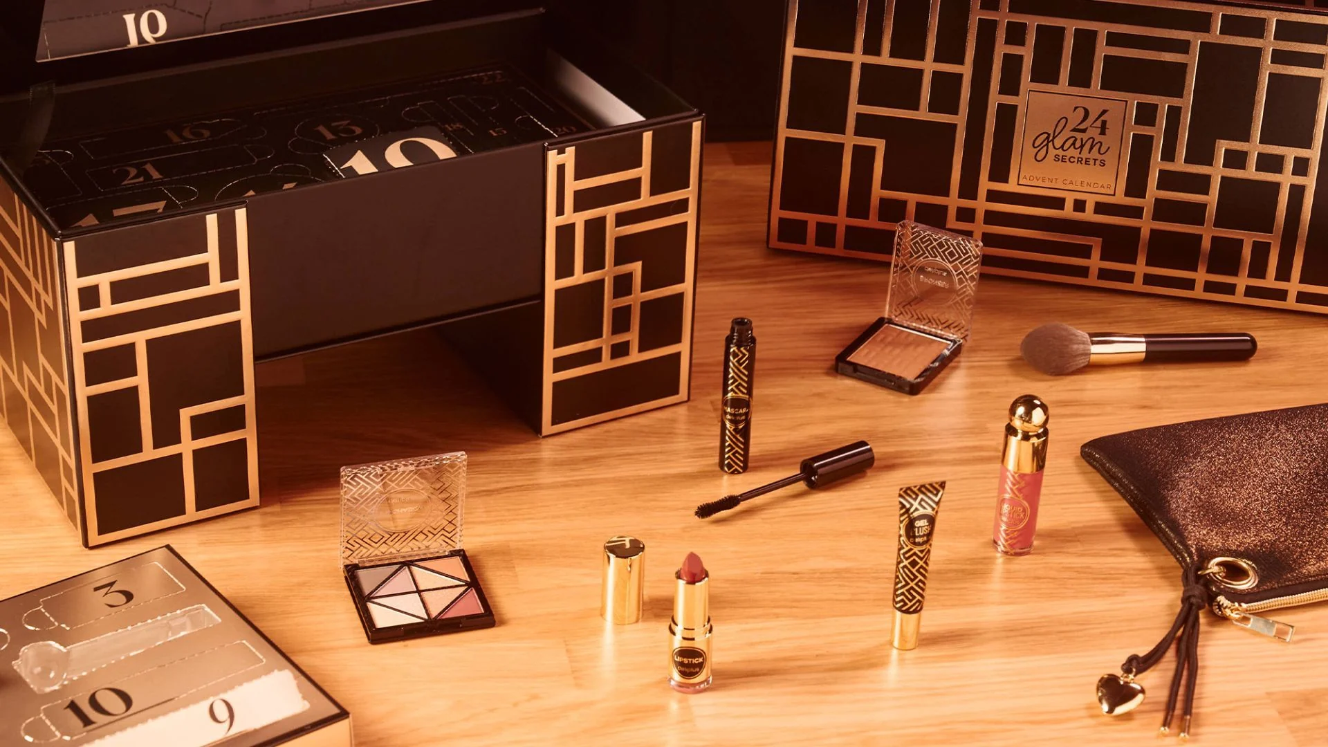 Caixa preta e dourada 24 Glam Secrets com compartimentos numerados, paleta, batom, máscara de pestanas, blush e bolsa sobre uma mesa.