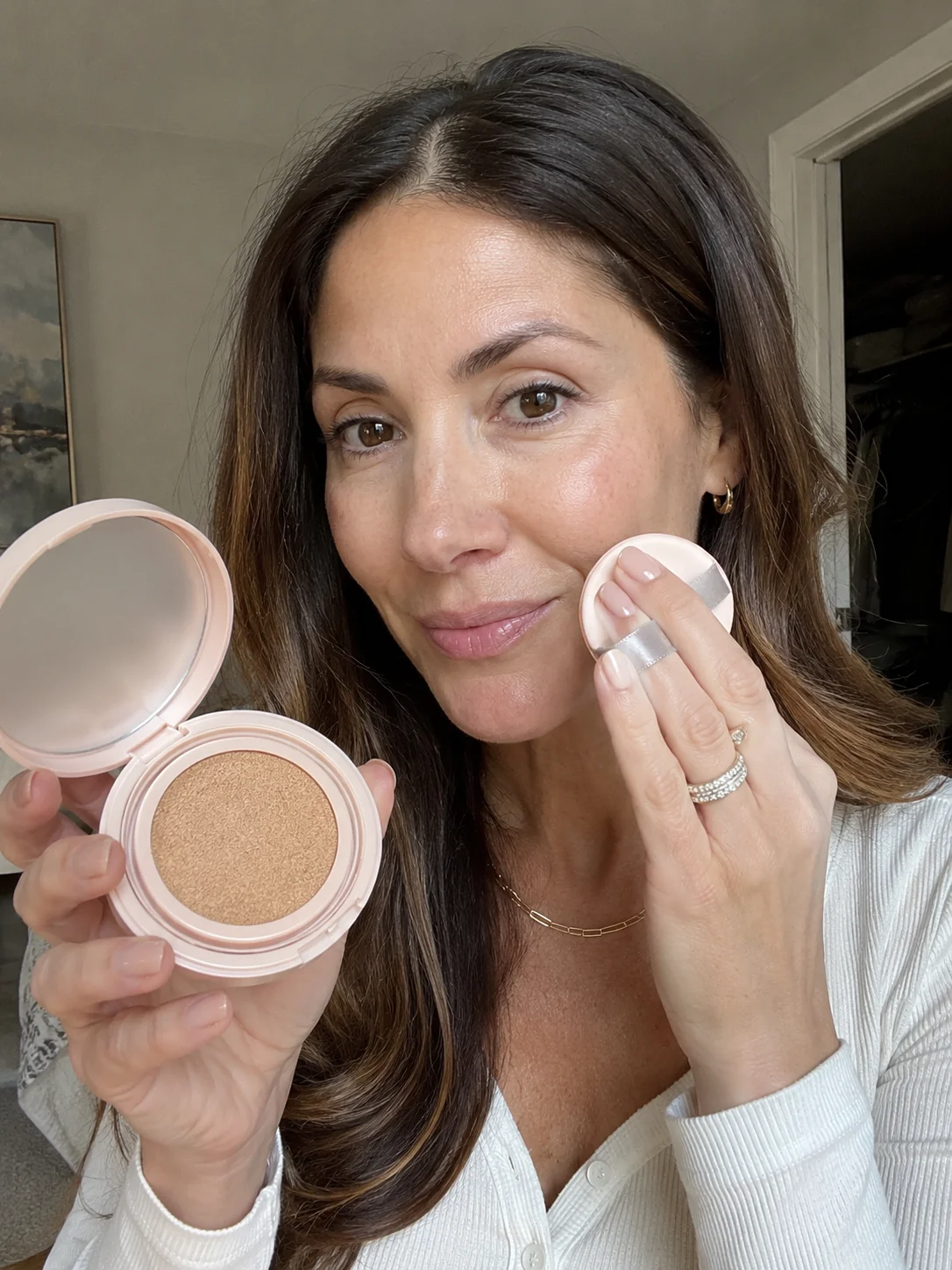 Mulher com pele madura a aplicar base cushion no rosto com puff junto a um compacto aberto.
