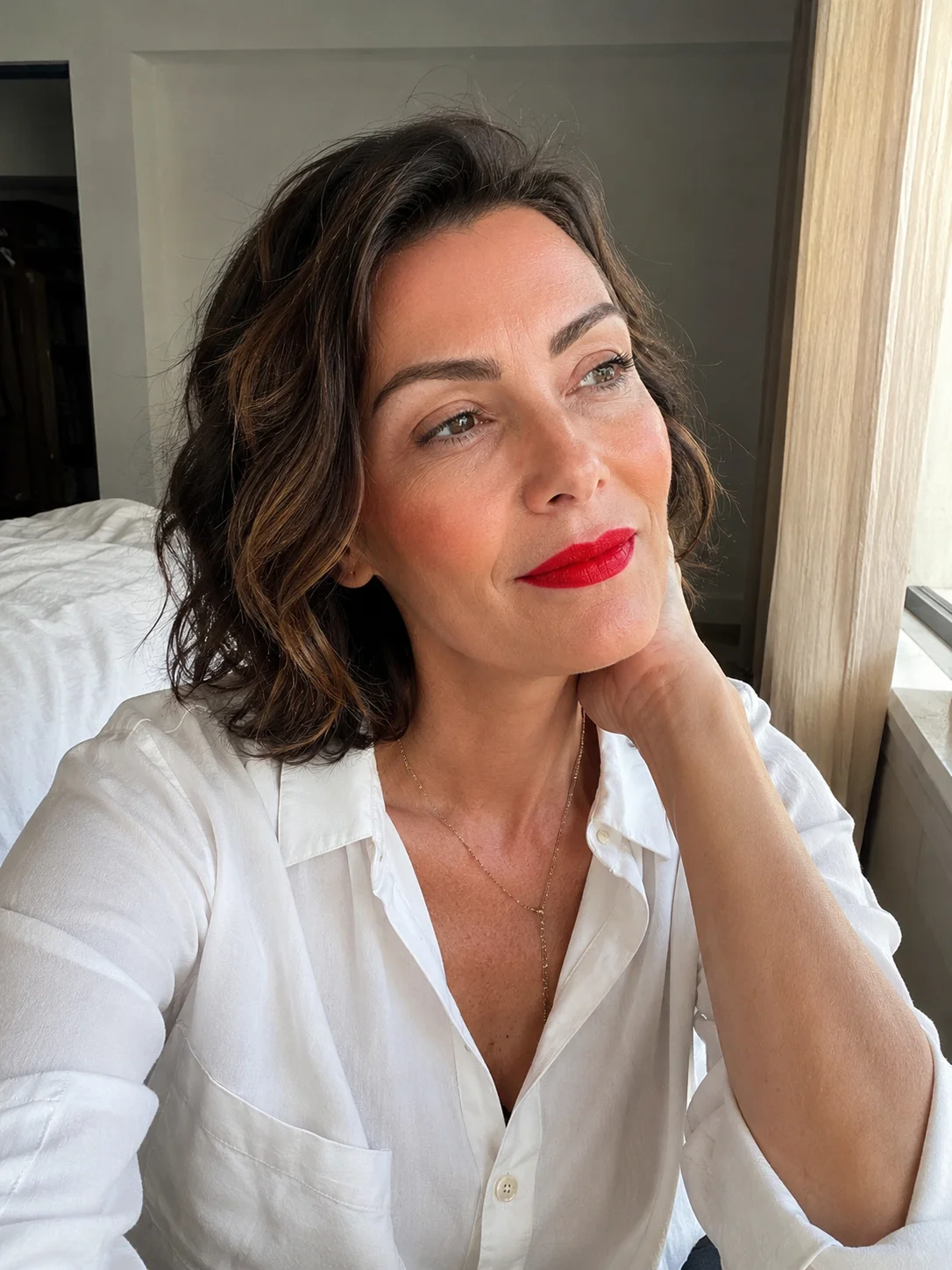 Mulher depois dos 50 com batom vermelho intenso e maquilhagem leve junto a uma janela