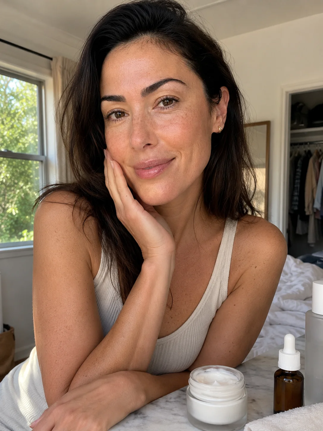 Mulher de meia-idade numa rotina de skincare em casa, com creme hidratante e sérum sobre a bancada e pele luminosa com textura natural visível.