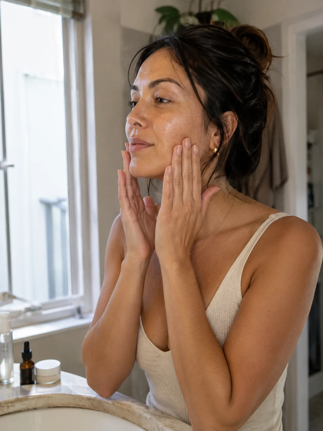 Mulher observa a pele do rosto junto a uma janela durante a rotina de skincare