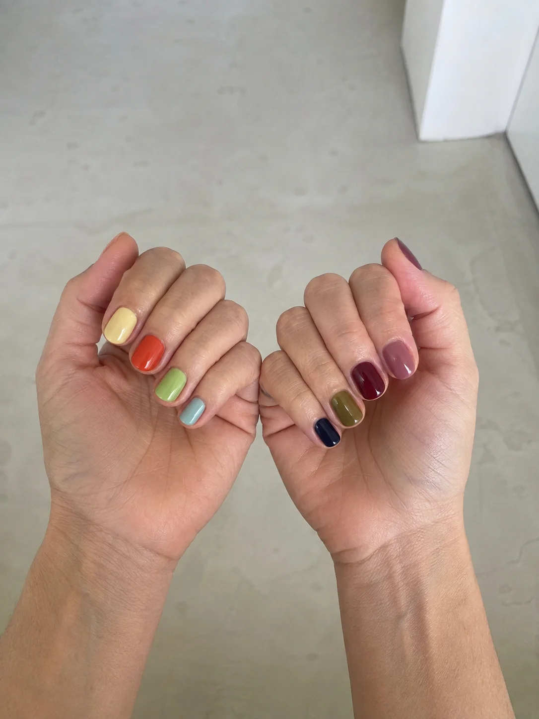 Duas mãos com unhas curtas em cores de verão e tons mais profundos de outono, incluindo amarelo suave, coral, verde, azul, bordô, oliva e mauve.