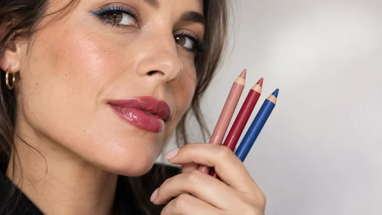 Mulher com lábios rosa brilhantes a segurar três lápis de maquilhagem em tons rosa, vermelho e azul.