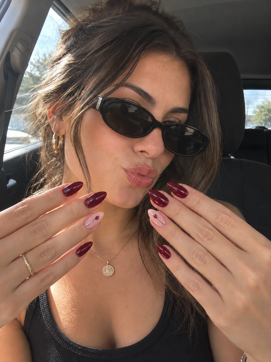 Mulher com óculos de sol a mostrar unhas amendoadas em bordô brilhante, com duas unhas de detalhe em rosa claro.